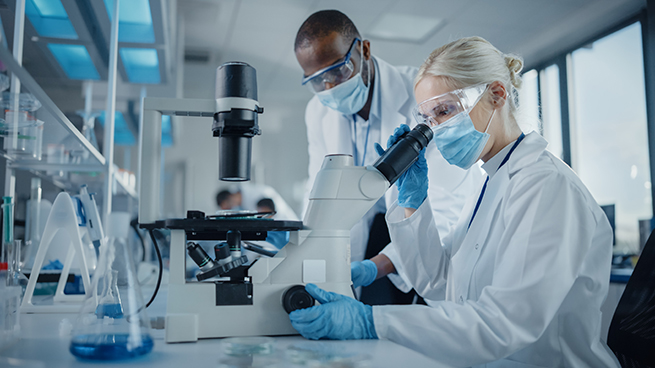 Modern Medical Research Laboratory: Two Scientists Wearing Face Masks Working Together Using Microscope, Analysing Samples, Talking. Advanced Scientific Lab for Medicine, Biotechnology.